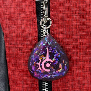 May include: A teardrop-shaped pendant with a silver clasp, featuring a dark purple resin filled with iridescent glitter. A pink emblem is embedded in the resin. The pendant hangs against a red textured fabric background.