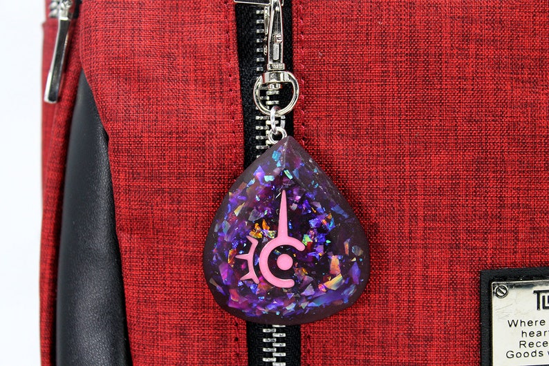 May include: A teardrop-shaped pendant with a silver clasp, featuring a dark purple resin filled with iridescent glitter. A pink emblem is embedded in the resin. The pendant hangs against a red textured fabric background.