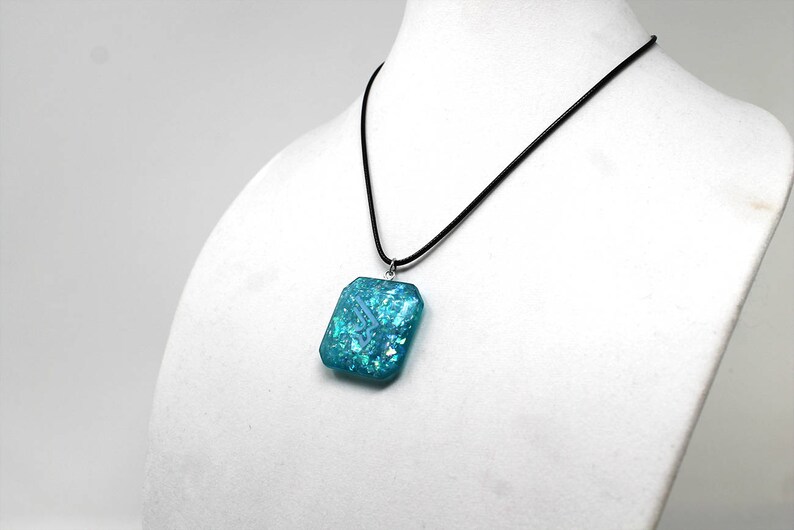 May include: A turquoise blue pendant necklace with a square-shaped pendant with iridescent glitter embedded in the resin. The pendant is hanging from a black cord.
