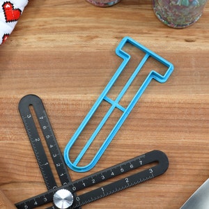 May include: A light blue plastic cookie cutter shaped like the letter T. The cutter is on a wooden surface with a ruler and other baking tools.