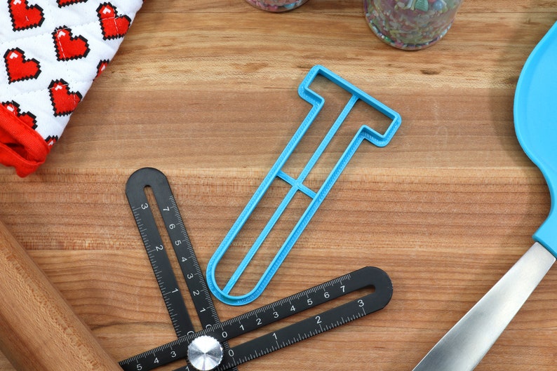 May include: A light blue plastic cookie cutter shaped like the letter T. The cutter is on a wooden surface with a ruler and other baking tools.