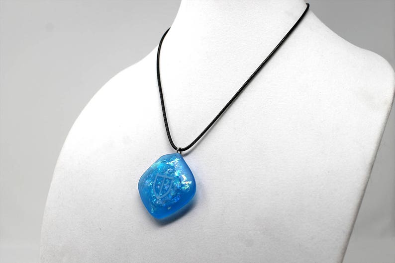 May include: A blue resin pendant with a white design hangs from a black cord necklace.