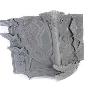 May include: A gray 3D printed book with intricate details, including a sword, feathers, and a chain. The book has a textured surface and appears to be a prop or decoration.