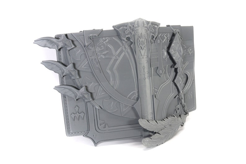May include: A gray 3D printed book with intricate details, including a sword, feathers, and a chain. The book has a textured surface and appears to be a prop or decoration.