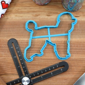 May include: A blue plastic cookie cutter shaped like a poodle dog. The cutter is on a wooden surface with a ruler and a rolling pin.
