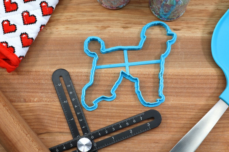 May include: A blue plastic cookie cutter shaped like a poodle dog. The cutter is on a wooden surface with a ruler and a rolling pin.