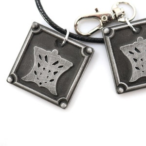 Puede incluir: Dos colgantes cuadrados plateados con un dise&ntilde;o decorativo, colgando de un cord&oacute;n negro con un cierre plateado.