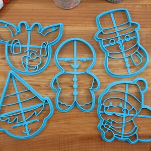 Op de afbeelding: Een set van vijf blauwe plastic kerstkoekjesvormen. De vormen zijn in de vorm van een rendier, een peperkoekman, een sneeuwpop, een elf en de kerstman.