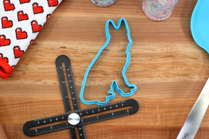 May include: Blue plastic cookie cutter shaped like a German Shepherd dog silhouette. The cutter is on a wooden surface with a ruler and other baking supplies.