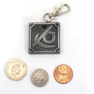 May include: A silver metal keychain with a stylized eye logo and a silver spring clasp. The keychain is next to three coins, one British pound coin and two US pennies.