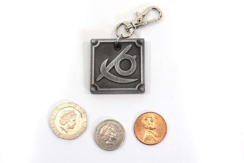 May include: A silver metal keychain with a stylized eye logo and a silver spring clasp. The keychain is next to three coins, one British pound coin and two US pennies.