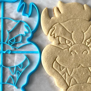 Op de afbeelding: Een blauwe plastic koekjesvormpje en een lichtbruine koek in de vorm van een cartoonmonster met een brede grijns.