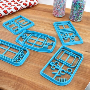 May include: Five blue plastic cookie cutters shaped like handheld video game consoles. The cutters are on a wooden surface.
