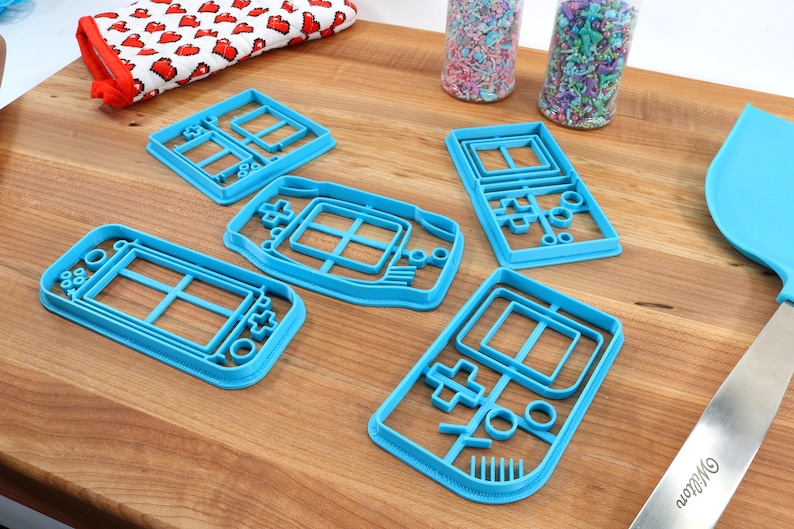 May include: Five blue plastic cookie cutters shaped like handheld video game consoles. The cutters are on a wooden surface.