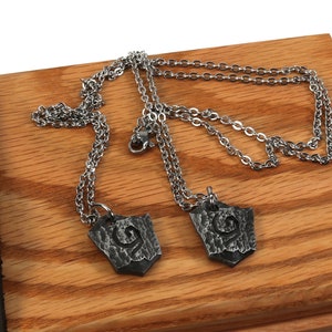 May include: Two silver chains with matching pendants. The pendants are silver and have a textured, shield-like shape with a design in the center.