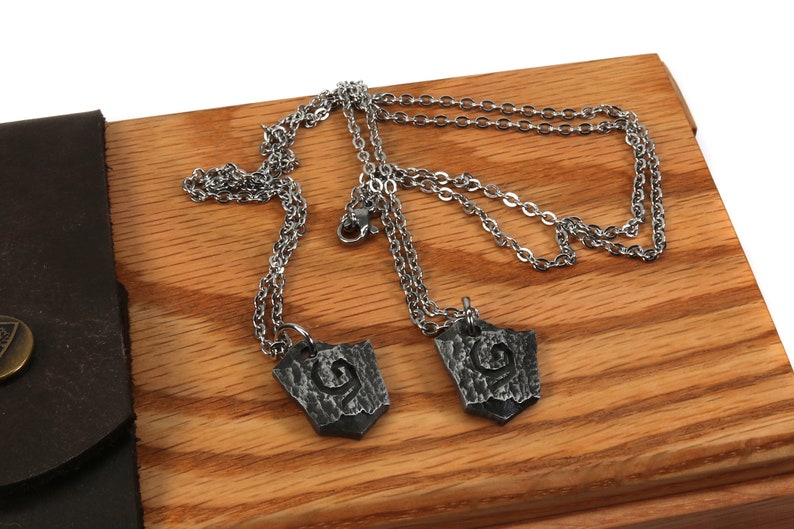May include: Two silver chains with matching pendants. The pendants are silver and have a textured, shield-like shape with a design in the center.