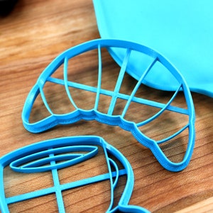 May include: Bright blue cookie cutters in various shapes, including a croissant and a coffee cup, are arranged on a wooden surface. A matching blue spatula is visible in the background. The cutters are made of plastic.