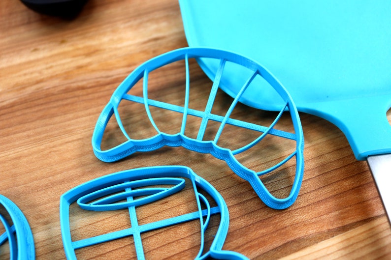 May include: Bright blue cookie cutters in various shapes, including a croissant and a coffee cup, are arranged on a wooden surface. A matching blue spatula is visible in the background. The cutters are made of plastic.