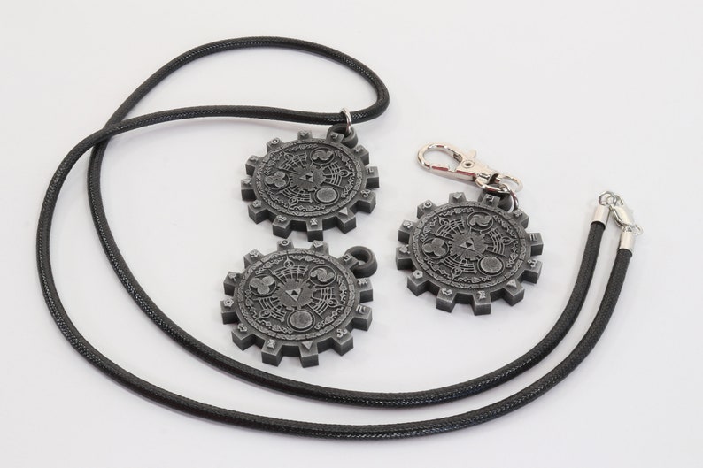 May include: Two silver-colored pendants with intricate designs, one on a black cord necklace and the other on a black cord keychain.