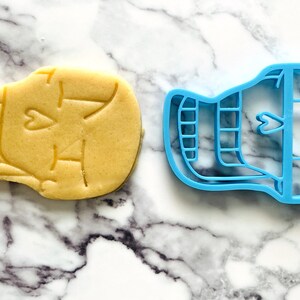 May include: A blue plastic cookie cutter shaped like a skull with a heart on the forehead. A sugar cookie is shown next to the cutter.