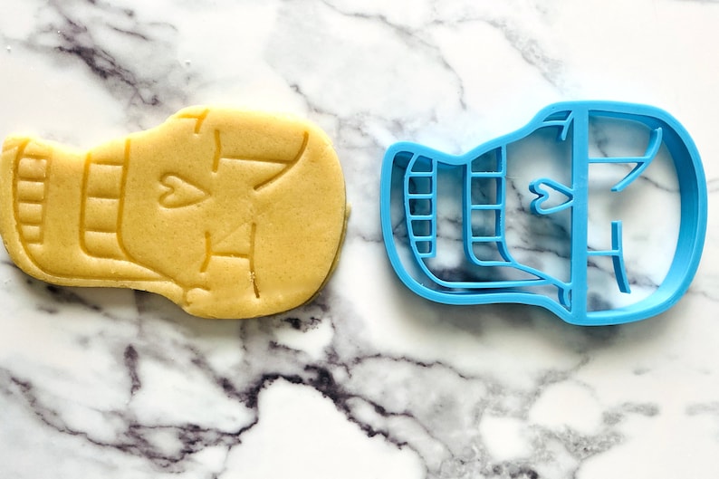 May include: A blue plastic cookie cutter shaped like a skull with a heart on the forehead. A sugar cookie is shown next to the cutter.
