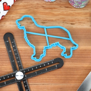 May include: A blue plastic cookie cutter shaped like a dog on a wooden surface with a black measuring tool and a blue spatula.