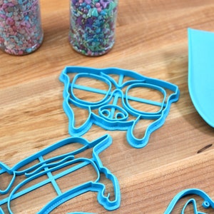 May include: A set of blue plastic cookie cutters shaped like dogs, including one with sunglasses. The cutters are on a wooden surface.