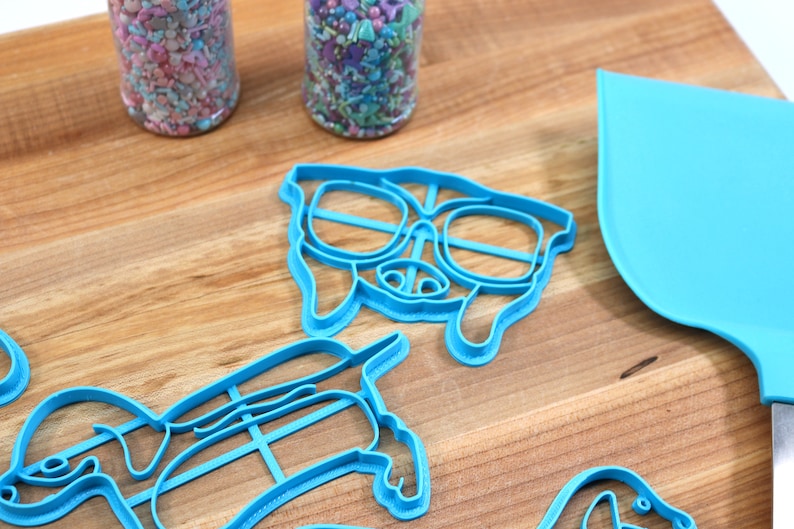 May include: A set of blue plastic cookie cutters shaped like dogs, including one with sunglasses. The cutters are on a wooden surface.