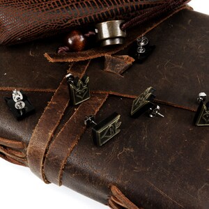 May include: Black crown-shaped stud earrings with a heart design, displayed on a brown leather surface. The earrings have a metallic finish and are set against a textured leather background, suggesting a vintage or handcrafted aesthetic.