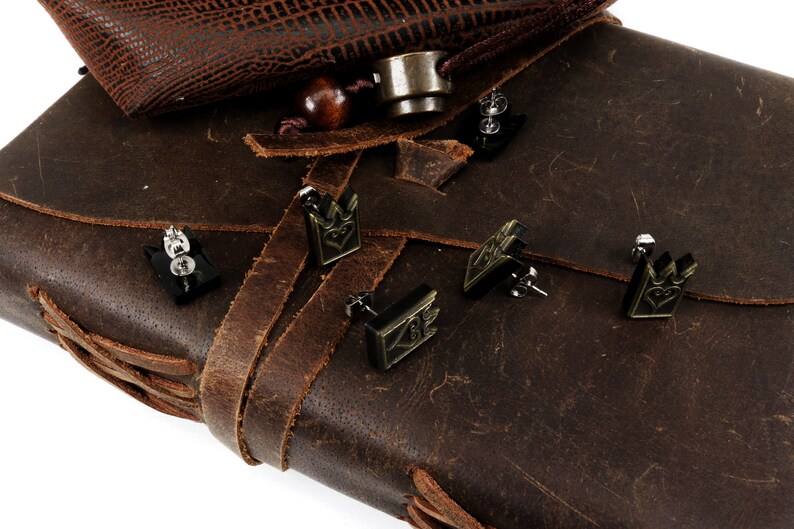 May include: Black crown-shaped stud earrings with a heart design, displayed on a brown leather surface. The earrings have a metallic finish and are set against a textured leather background, suggesting a vintage or handcrafted aesthetic.