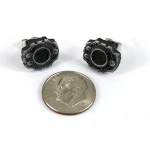 May include: A pair of silver stud earrings with a black, circular design. The design features a raised, textured border.