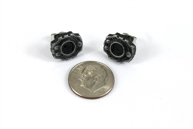 May include: A pair of silver stud earrings with a black, circular design. The design features a raised, textured border.
