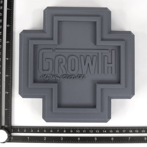 May include: A gray, 3D printed cross-shaped object with the word "GROWTH" and the phrase "My Way TOLD" embossed on the front.