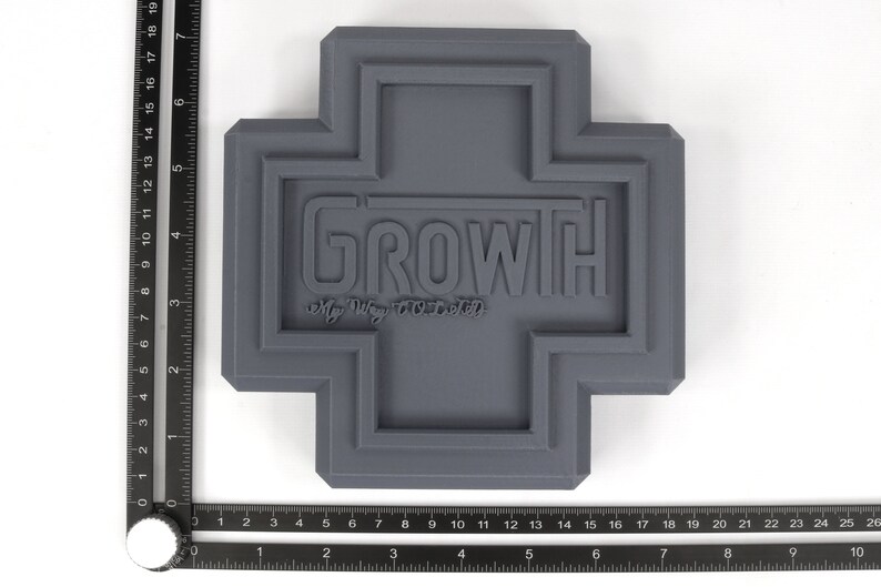 May include: A gray, 3D printed cross-shaped object with the word "GROWTH" and the phrase "My Way TOLD" embossed on the front.