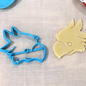 May include: A blue plastic cookie cutter shaped like a dragon's head and a light brown cookie in the shape of a dragon's head.