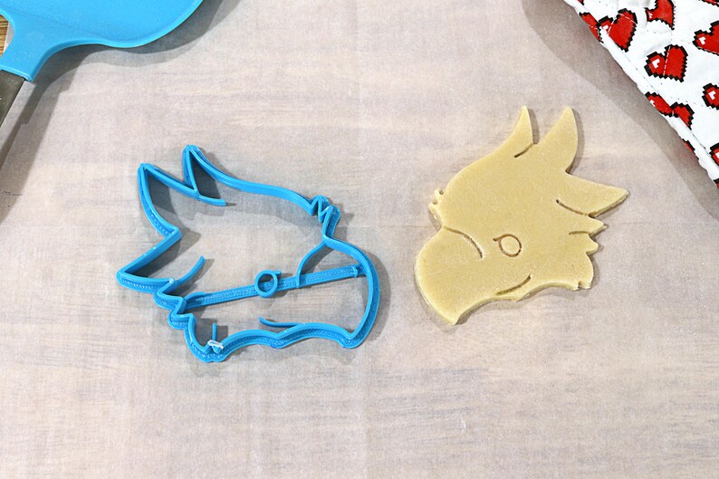 May include: A blue plastic cookie cutter shaped like a dragon's head and a light brown cookie in the shape of a dragon's head.