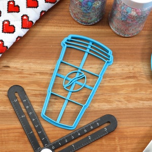 May include: A blue plastic cookie cutter shaped like a coffee cup with a coffee bean logo. The cutter is on a wooden surface with other baking supplies.