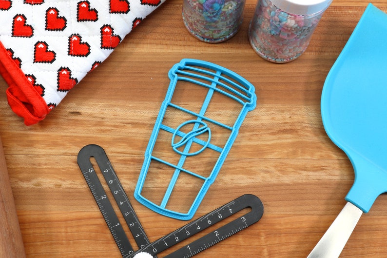 May include: A blue plastic cookie cutter shaped like a coffee cup with a coffee bean logo. The cutter is on a wooden surface with other baking supplies.