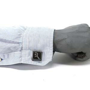 May include: A close-up of a gray metal cufflink with a magnifying glass design. The cufflink is on a blue and white striped shirt cuff. The shirt cuff is buttoned and the background is white.