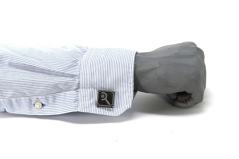 May include: A close-up of a gray metal cufflink with a magnifying glass design. The cufflink is on a blue and white striped shirt cuff. The shirt cuff is buttoned and the background is white.