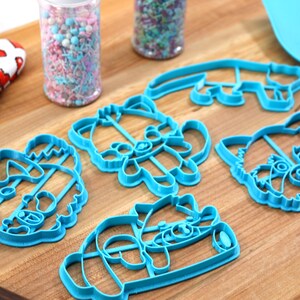May include: Set of five blue plastic cookie cutters shaped like raccoons. The cutters are on a wooden surface.