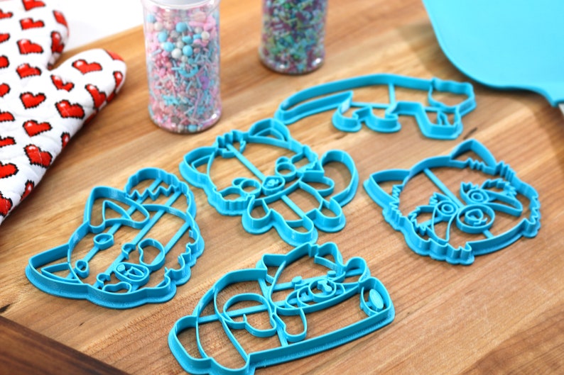 May include: Set of five blue plastic cookie cutters shaped like raccoons. The cutters are on a wooden surface.