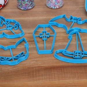 May include: Five blue plastic Halloween cookie cutters on a wooden surface. The cutters are shaped like a bat, a witch's hat, a coffin, a ghost, and a cat.