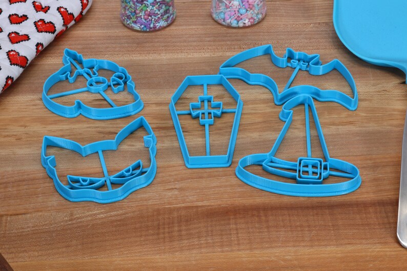 May include: Five blue plastic Halloween cookie cutters on a wooden surface. The cutters are shaped like a bat, a witch's hat, a coffin, a ghost, and a cat.