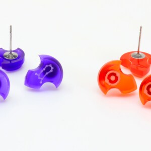 May include: Three pairs of earrings, two purple and two orange, with a silver post. The purple earrings have a white Gemini zodiac symbol. The orange earrings have a white circle in the center.