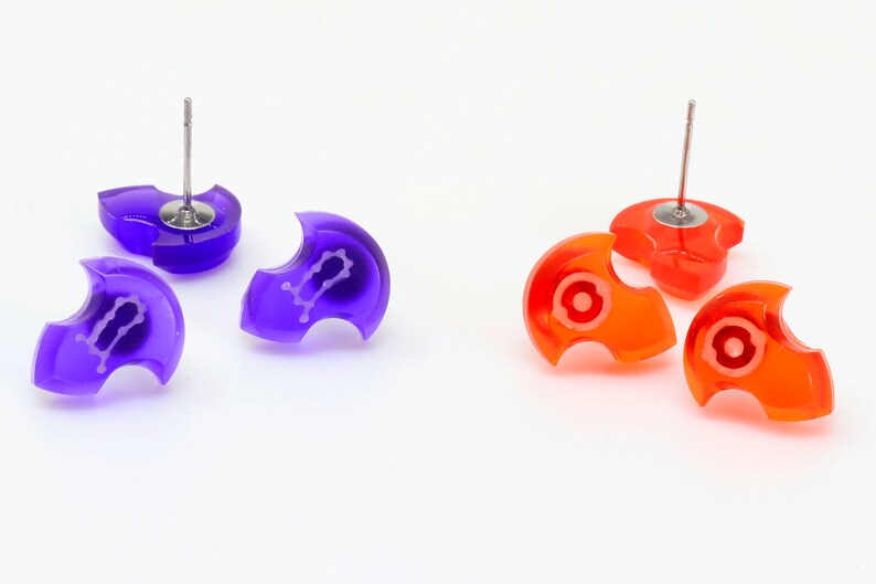 May include: Three pairs of earrings, two purple and two orange, with a silver post. The purple earrings have a white Gemini zodiac symbol. The orange earrings have a white circle in the center.