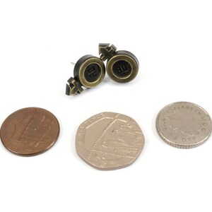 Puede incluir: Un par de pendientes dorados y negros con un s&iacute;mbolo de signo masculino y femenino. Los pendientes se muestran sobre una superficie blanca con tres monedas.