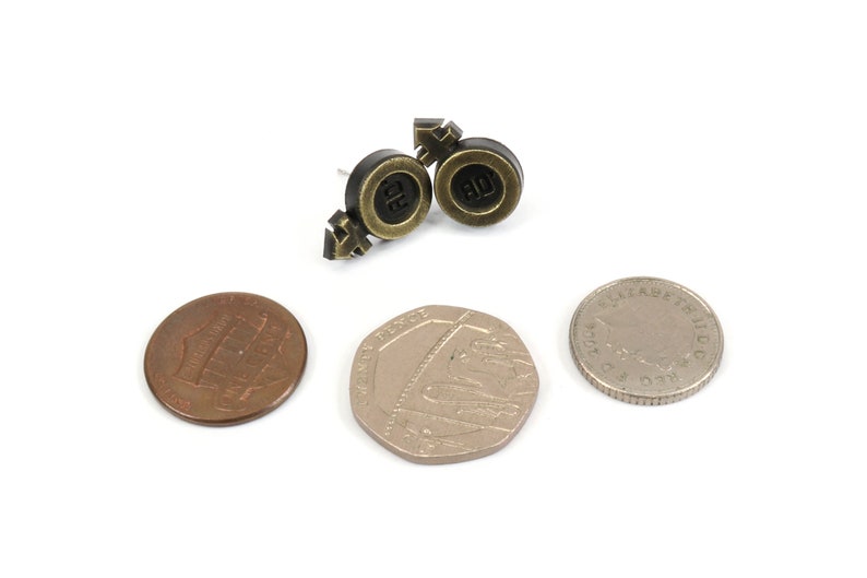 Puede incluir: Un par de pendientes dorados y negros con un s&iacute;mbolo de signo masculino y femenino. Los pendientes se muestran sobre una superficie blanca con tres monedas.