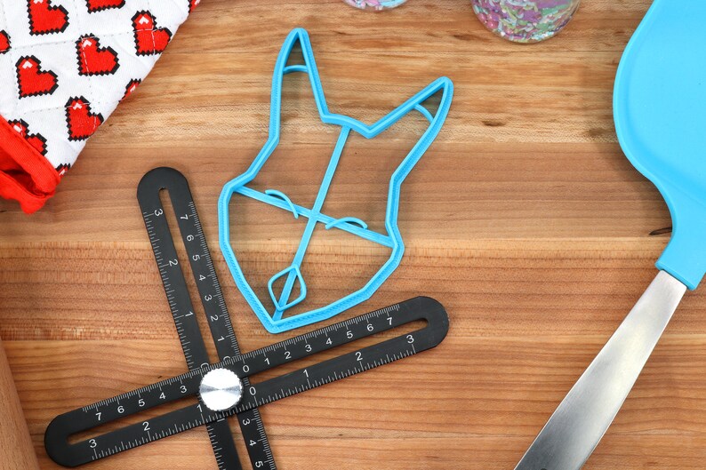 May include: A blue plastic cookie cutter shaped like a dog's head. The cutter is on a wooden surface with a black ruler.