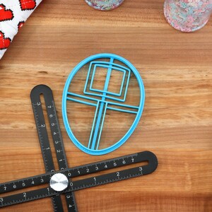 May include: A blue plastic cookie cutter shaped like a circle with a square and lines inside. The cutter is on a wooden surface with a black ruler and a white and red fabric with hearts.